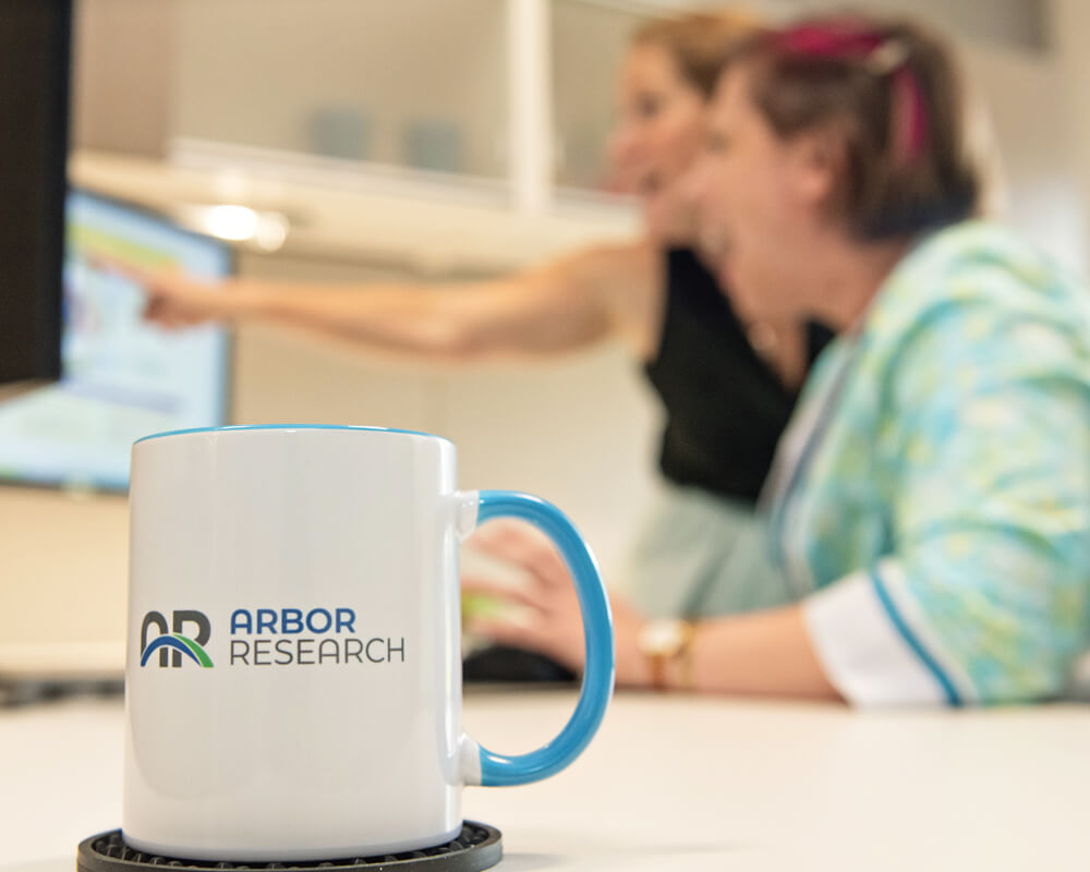 In the background, a woman in an office setting points out a detail on the screen to her coworker. In the foreground is an Arbor Research branded coffee mug.